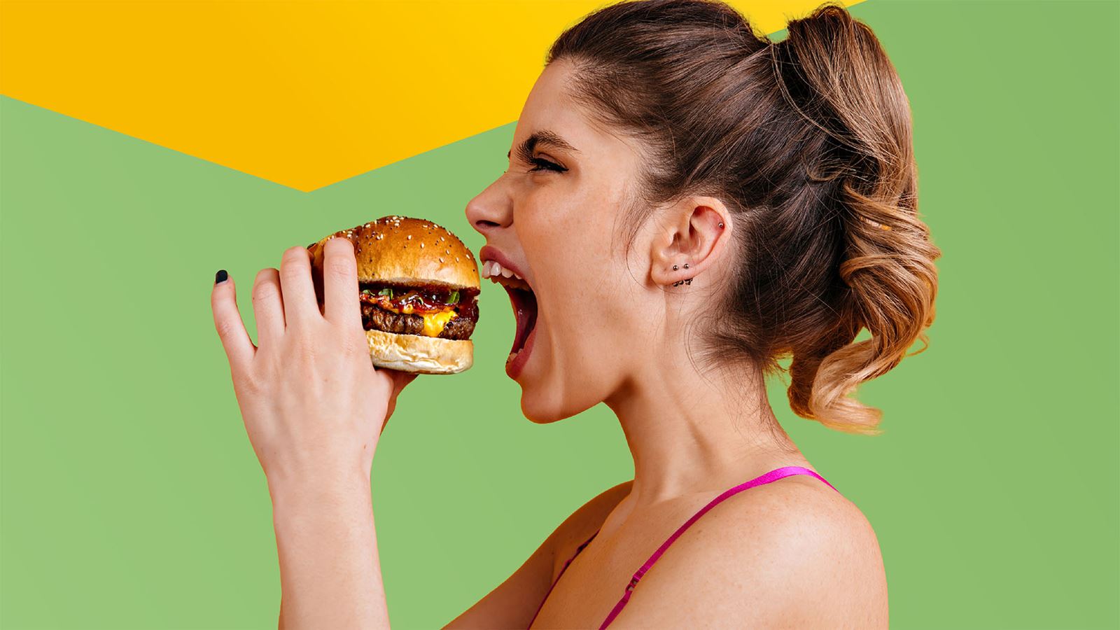 A young woman bites a juicy burger with anticipation (with yellow design element above)