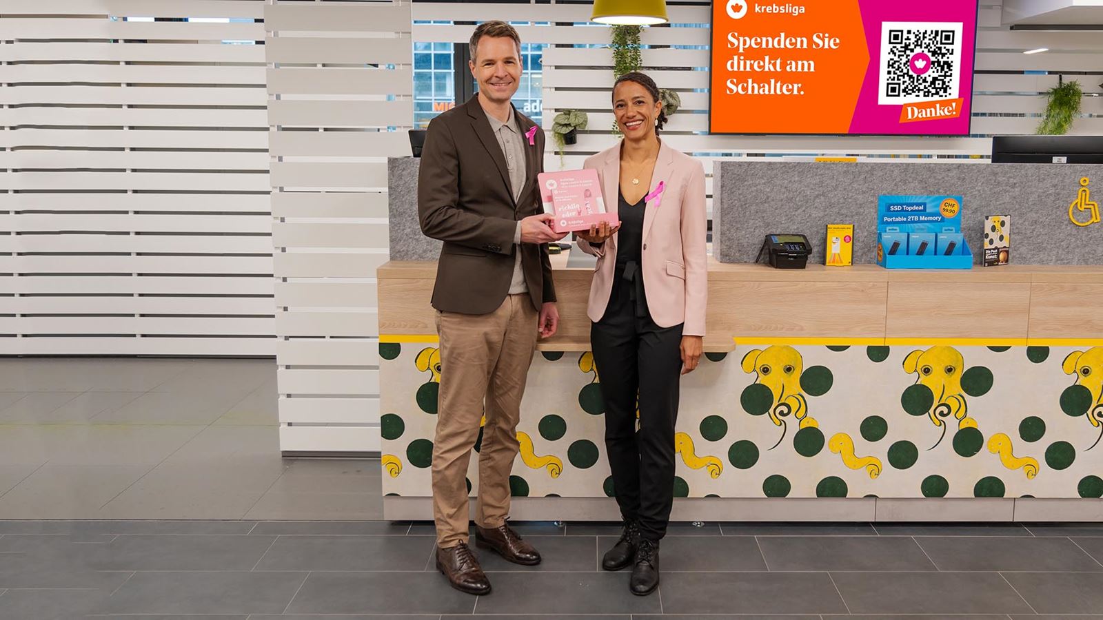 Oliver Egger, Head of Media & Advertising Market at Swiss Post Advertising and Mirjam Lämmle, CEO of the Swiss Cancer League, present the pink awareness ribbons at a Swiss Post branch.