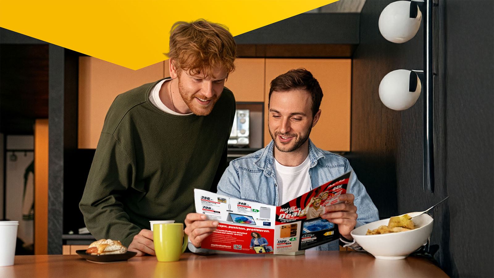 Two young men study a MediaMarkt product catalogue at the breakfast table (with a yellow design element above)