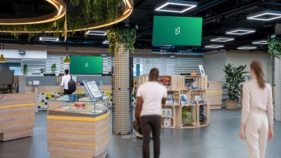 Innenansicht eines modernen Verkaufs- oder Aufenthaltsbereichs mit mehreren digitalen Screens an der Decke; im Vordergrund sind vorbeigehende Personen und verschiedene Präsentationsflächen zu sehen.