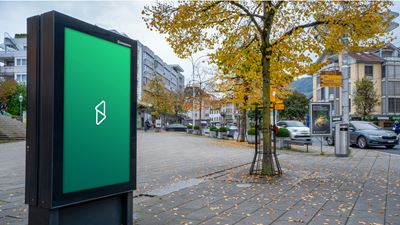 Digitaler Werbescreen im öffentlichen Raum an einer Strasse oder in einem Stadtzentrum; im Hintergrund sind Bäume, Gebäude und vorbeifahrende Autos zu sehen.